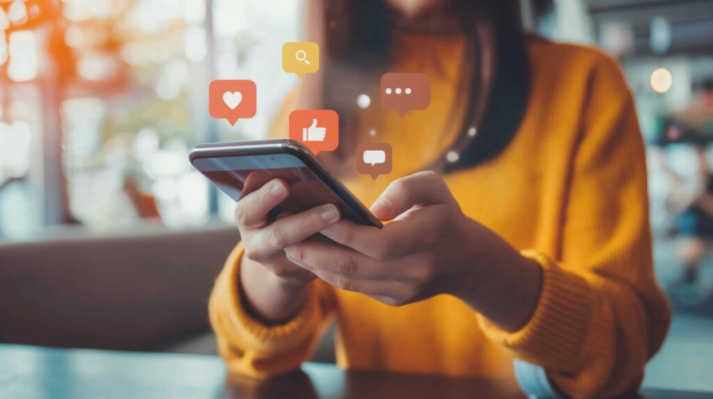 Eine Frau im gelben Pullover schaut auf ihr Smartphone. Aus diesem kommen Social Media Icons herausgeflogen.